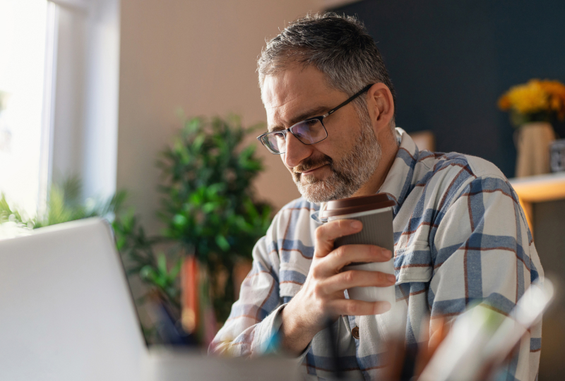 Man smiling as he holds a cup of coffee in his right hand and is looking towards his laptop screen.