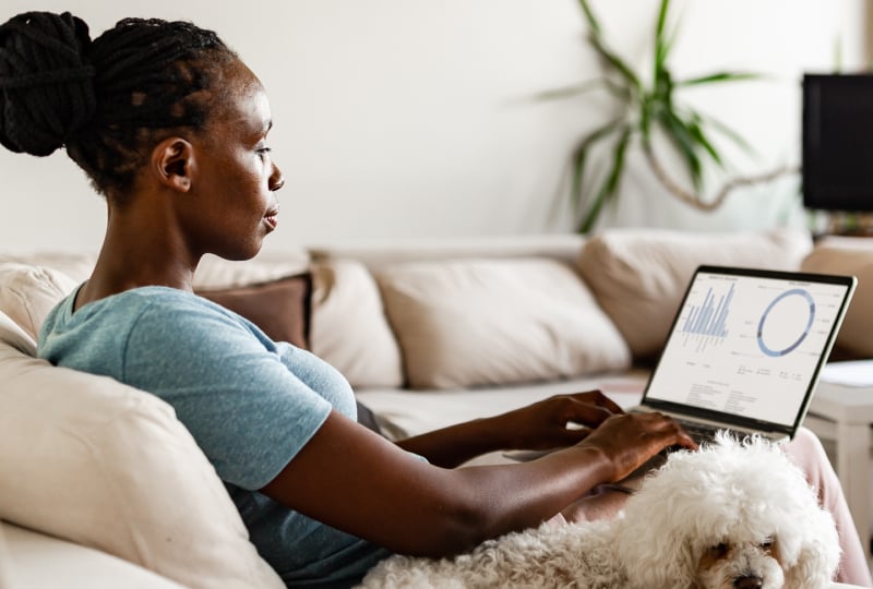 Woman sitting on a couch with her dog sitting next to her as she works on a laptop.