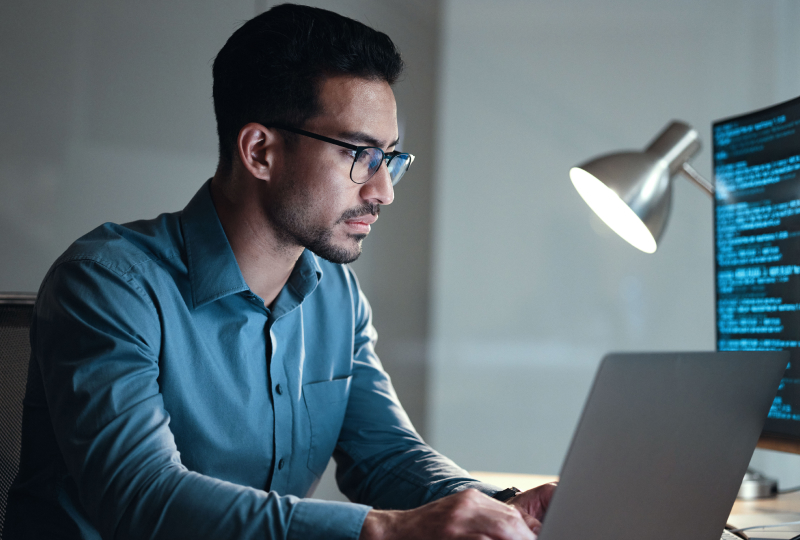 Man sitting at a desk with a lamp shining towards him while he works on a laptop.