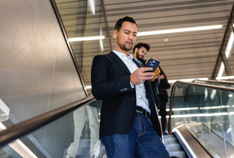 A professional man in a suit rides an escalator down while looking at his cell phone