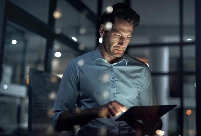 Man in a dimly lit office standing and interacting with a tablet in his hands.