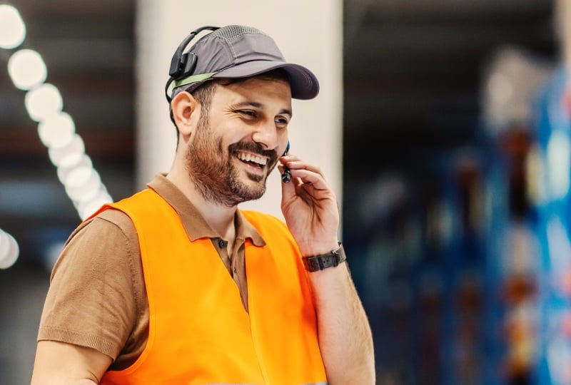 Smiling man wearing a orange reflective vest interacting with the mouthpiece of a mobile headset he is wearing.